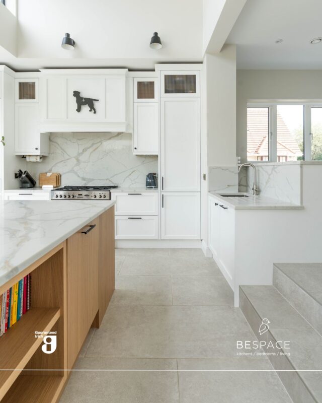 A layered space designed with flow and function in mind 💭

This pristine white kitchen features elegant marble worktops and splash back, with an island set at a level below. The warm oak tones and black handles add contrast and character, tying in seamlessly with the silver and black oven 🖤 

Up a few steps, the main cabinetry and extra countertop space completes this kitchen. Upgrade your space today | Link in bio